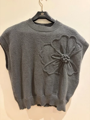 Grå stickad väst med blomdetalj H&M - Säljer en grå stickad väst från H&M med en stor, upphöjd blomdetalj på framsidan. Västen har ribbade kanter vid hals, ärmhål och nederkant. Perfekt att styla över en skjorta eller långärmad tröja för en trendig lager-på-lager-look.