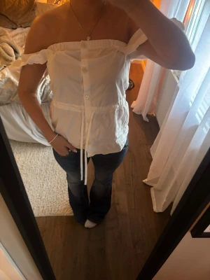 Vit offshoulder blus med knappar - Säljer en söt vit offshoulder blus med knappar framtill och knytband i midjan. Blusen har volangkant upptill och är ärmlös, perfekt för varma dagar. Materialet känns lätt och luftigt, vilket gör den superbekväm att bära.