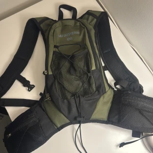 Grön vätskesystem ryggsäck 12L - Säljer en grön och svart ryggsäck från VÄTSKESYSTEM, modell 1201, med plats för vätskebehållare och slang. Ryggsäcken har flera fack med dragkedja, meshfickor på sidorna och justerbara remmar. Perfekt för träning eller äventyr utomhus. OBS!! Saknar påse för vätska!