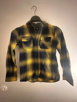 Rutig gul och blå skjorta från Vans - Snygg rutig skjorta från Vans i gult och mörkblått. Skjortan har två bröstfickor med knappar och klassisk krage. Tillverkad i mjuk bomullsflanell och har långa ärmar. Juniorstorlek M, motsv ca 160