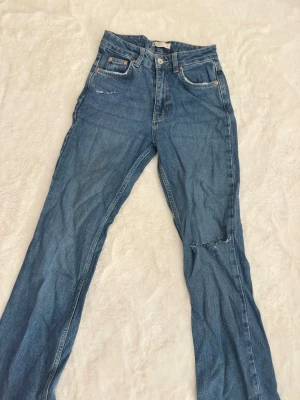Blå mid waist jeans  - Klassiska blå jeans med medium midja från Gina tricot . Modellen har raka ben och 4 fickor, samt slitningar och hål på ena benet för en avslappnad look. Jeansen är tillverkade i tåligt denimtyg och har kopparfärgade nitar och knapp.