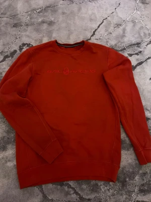 Röd Sail Racing sweatshirt - Säljer en röd sweatshirt från Sail Racing med broderad logga på bröstet. Tröjan har rund hals, långa ärmar och är gjord i mjuk bomull. Perfekt för dig som gillar stilrena plagg med enkel design.