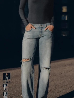 Ljusblå låga jeans med slitningar - Säljer ett par ljusblå jeans med breda ben och slitna detaljer vid knäna. Jeansen har klassisk femficksdesign, låg midja och är tillverkade i jeansmaterial. Perfekta för en avslappnad och trendig look.