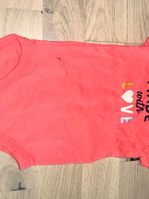 Rosa body från Carter's med tryck Carter's korallfärgad body, storlek 6M - Söt rosa body från Carter's i mjuk bomull. Framsidan har ett färgglatt tryck med texten 'Made with Love' där bokstäverna är i olika färger och ett hjärta ersätter O:et. Perfekt för små bebisar som vill ha något gulligt och bekvämt.Söt korallfärgad body med korta ärmar och texten "Made with Love".