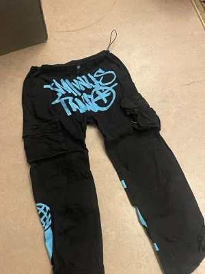 Svarta cargopants med blått tryck - Unika svarta cargopants med stora blå graffiti-prints fram och bak. Byxorna har flera fickor på sidorna och justerbara benslut med snörning. Tillverkade i bomull för en skön och loose passform. Perfekta för dig som vill sticka ut med streetwear-vibe.