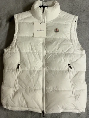 Vit dunväst från Moncler - Säljer en stilren vit dunväst från Moncler med hög krage och två fickor med dragkedja. Västen har quiltad design och klassisk Moncler-logga på bröstet. Perfekt att slänga över en hoodie eller tröja för extra värme.