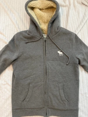 Fodrad Abercrombie & fitch grå zip up hoodie - Vintage gråfodrad unisex Abercrombie & Fitch huvtröja med dragkedja. Herrstorlek S! Fodrad inuti och med den vita Abercrombie & Fitch älglogotypen på bröstet. Mycket bra skick förutom 2 mycket små defekter som syns på bilderna! Fodret inuti är också lite missfärgat!!