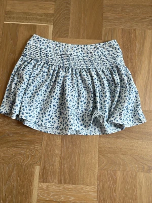 Blommig kortkjol från Gina Tricot - Supersöt kortkjol från Gina Tricot med blått blommönster på vit botten. Kjolen har smock i midjan för en bekväm passform och ett lätt, luftigt tyg som passar perfekt till varma dagar. Perfekt att matcha med en enkel topp eller hoodie.