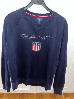Marinblå sweatshirt från GANT  - Säljer en marinblå sweatshirt från GANT i storlek XL. Tröjan har broderad logga och sköld med stjärnor och ränder på bröstet. Klassisk rund halsringning och långa ärmar. Tillverkad i mjuk bomull för skön känsla.