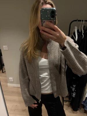 Beige stickad kofta från 157 - Mysig beige stickad kofta från 157 med dragkedja framtill och ribbad struktur. Koftan har långärmad passform och hög krage, perfekt för lager på lager under kyliga dagar.