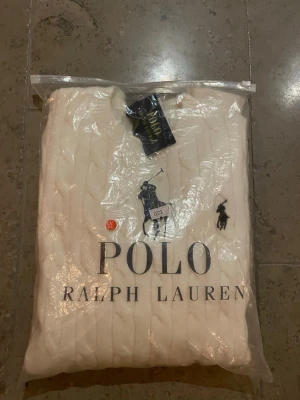 Vit kabelstickad tröja Polo Ralph Lauren - Helt ny, aldrig använd, kabelstickad vit tröja från Polo Ralph Lauren med klassisk rund hals och broderad logga på bröstet. Tröjan har ribbade muddar och är perfekt för dig som gillar en clean och tidlös stil. Materialet är mjukt och känns lyxigt mot huden.