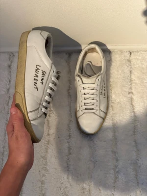 Vita Saint Laurent skor - Exklusiva Saint Laurent  sneakers i vit färg med ikonisk graffiti-logga. Tidlös modell i högkvalitativt italienskt läder – passar lika bra till vardag som till uppklädda looks.  ✔️ Märke: Saint Laurent Paris✔️ Skick: Mycket bra begagnat skick✔️ Anmärkning: Endast lätt smuts på sulan✔️ Detaljer: Originalkort medföljer  Skorna är väl omhändertagna och har lång livslängd kvar. Nypris flera tusen kronor – perfekt för dig som vill ha äkta design till bättre pris.  📦 skickar snabbt