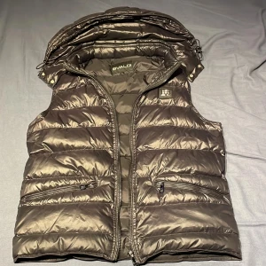 BVALDI Väst - En varm väst med två fickor på utsidorna  om västen och 1 ficka på insidan. Man kan även ta av och sätta på luvan. Passar dig som har M 👍