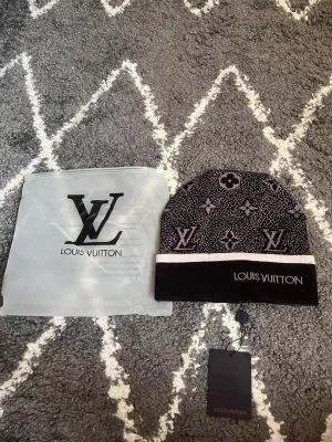 L-mössa  -  Exklusiv mössa från L i  grått och svart med klassiskt L-monogram och blommönster. Mössan har en svart uppvikt kant med vitta rand och broderad L-logga framtill. Tillverkad i mjukt stickat material, perfekt för kyliga dagar.