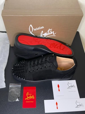 Christian Louboutin svarta sneakers med nitar - Säljer ett par svarta sneakers från Christian Louboutin i mocka med ikonisk röd sula och svarta nitar över tån. Skorna har snörning och en rund tå, vilket ger en edgy och lyxig vibe. Perfekta för dig som vill sticka ut med exklusiva detaljer.