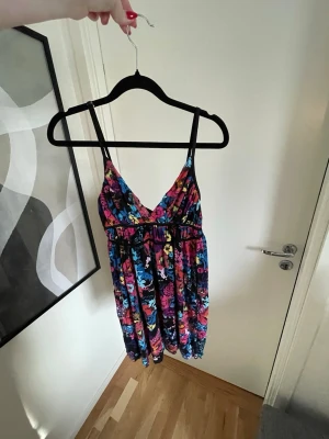 Färgglad blommig sommarklänning - En färgglad blommig klänning med smala axelband och v-ringning. Klänningen har svart botten och mönster i rosa, blått, gult och lila