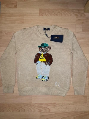 Beige stickad tröja Polo Ralph Lauren - Beige stickad tröja från Polo Ralph Lauren med ikonisk björn på bröstet. Tröjan har rund halsringning, ribbade muddar och RL-broderi nedtill. Perfekt för dig som vill ha en klassisk men lekfull look.