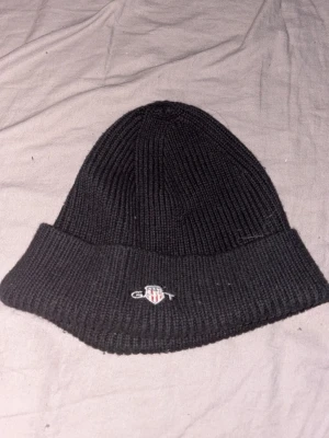 Svart ribbstickad mössa från Gant - Svart ribbstickad mössa från Gant med uppvikt kant och broderad logga framtill. Klassisk och enkel design som passar till allt. Perfekt för kyliga dagar och ger en clean look.