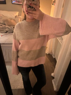 Randig rosa och beige stickad tröja - Mysig stickad tröja med breda ränder i ljusrosa och beige. Tröjan har lång ärm med lite vidare passform och rund halsringning. 