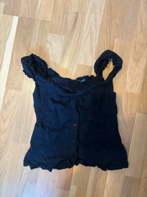 Svart blus från Brandy Melville - Säljer en svart blus från Brandy Melville med knappar framtill och volangdetaljer vid axelbanden