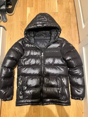 Svart pufferjacka från Alessandro  - Säljer en svart pufferjacka från Alessandro. Blank finish med huva och dragkedja framtill. Varm och bekväm, perfekt för höst/vinter.
