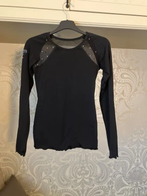 Svart Bolero Ice Dance topp med strass - Supersnygg svart långärmad topp från Bolero Ice Dance i stretchigt material. Dekorerad med små strasstenar på rygg och framtill och ett hjärta i strass på ärmen. Meshdetaljer vid axlarna och färgglada stenar på ena ärmen. Nypris ca 800kr. Saknar någon enstaka strass-sten.