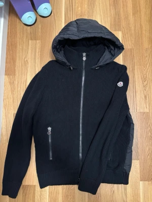 Svart stickad pufferjacka från Moncler - Säljer en svart pufferjacka från Moncler med stickade ärmar och sidor, samt quiltad huva och rygg. Jackan har dragkedja framtill och två fickor med dragkedja. Klassisk Moncler-logga på ärmen. Perfekt för kyliga dagar.