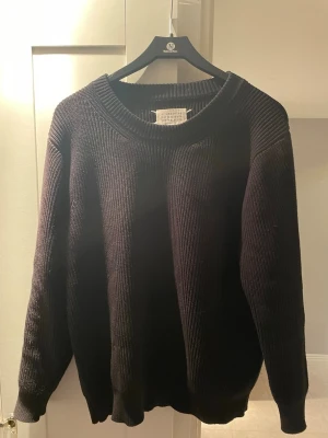 Masion Margiela Knit - Säljer en svart stickad tröja från Maison Margiela. Tröjan har en bred ribbad rundhals, långa ärmar och den ikoniska Margiela-lappen i nacken. Perfekt oversized fit och tillverkad i Italien. Grym basic för garderoben! Storlek S men sitter som en M beroende på om du vill ha den lite tightare eller föredrar en loose fit. 