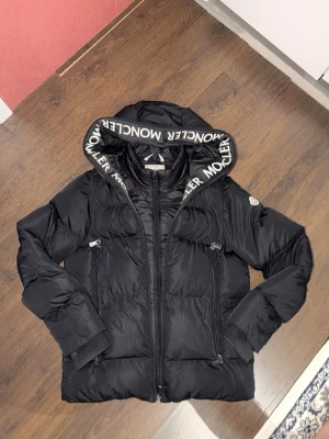 Svart Moncler pufferjacka med huva - Säljer en svart pufferjacka från Moncler med stor huva och logga på kanten. Jackan har dragkedja framtill, två sidofickor med dragkedja och ribbade muddar vid ärmsluten. Insidan har coola Moncler-etiketter och serier. Perfekt för kalla dagar och riktigt snygg streetstil.