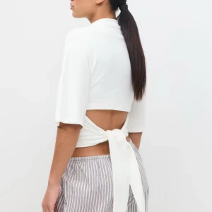 Vit topp med knytning i ryggen - Snygg vit topp med halvpolokrage och trekvartsärmar. Toppen har en öppen rygg med breda band som knyts i midjan för en cool och trendig look. Perfekt att styla med höga jeans eller kjol för en fräsch vibe.
