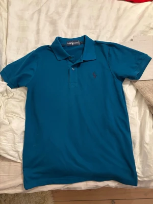 Turkose/blå pikétröja från Ralph Lauren - Säljer en klassisk blå/turkose pikétröja från Ralph Lauren i storlek M. Tröjan har korta ärmar, krage och knappar framtill samt den ikoniska broderade loggan på bröstet. Perfekt skick!
