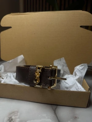 Mörkbrunt YSL bälte i skinn - Snyggt mörkbrunt bälte från YSL i äkta skinn med guldfärgat spänne och ikonisk YSL-logga framtill. Perfekt accessoar för att lyfta vilken outfit som helst med en lyxig touch. Bältet har klassisk design och exklusiva detaljer.