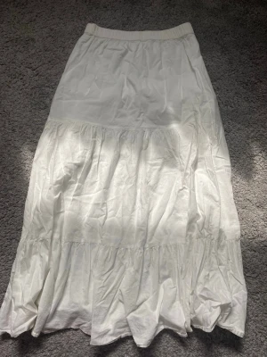 Vit långkjol med volang - Säljer en vit långkjol med resår i midjan och flera volangpartier som ger en flowig och luftig känsla. Kjolen är tillverkad i ett lätt bomullstyg och passar perfekt för varma dagar. Enkel och stilren design som är lätt att matcha. En liten fläck längs ner på baksidan. Storlek 158