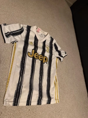 Juventus matchtröja KULUSEVSKI 44 barn - Juventus hemmatröja från Adidas med KULUSEVSKI och nummer 44 på ryggen. Tröjan är vit med svarta ränder och gulddetaljer, tillverkad i lätt och ventilerande AEROREADY-material. Klassisk V-ringning och Jeep-logga på bröstet.