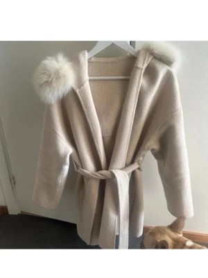 Beige kappa med pälskantad huva - Snygg beige kappa med stor huva kantad av fluffig päls. Jackan har bälte i midjan för att knyta. Säljer då jag inte har fått nån användning av den på länge. Pris kan diskuteras vid snabbt köp🥰