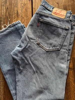 Levi's 501 ljusblå jeans med slitningar - Klassiska Levi's 501 jeans i ljusblå tvätt med raka ben och slitna detaljer på knäna. Jeansen har fem fickor, knappgylf och den ikoniska läderpatchen bak i midjan. Perfekt för dig som gillar en avslappnad och vintage-inspirerad look.