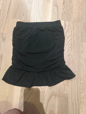 Glittrig svart kjol från Kappahl - Supersnygg svart kjol från Kappahl med glittriga detaljer och volang längst ner. Kjolen är kort och har en figurnära passform som ger en trendig look. Perfekt för dig som vill sticka ut lite extra!