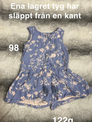 Blommig blå byxdress med tofsar - Söt blå byxdress med ljusa blommor och dekorativa tofsar i midjan. Ärmlös modell med resår i midjan och korta ben. Perfekt för varma dagar och enkel att ta på. 🌸 Ena lagret tyg har släppt från en kant. Ej hål rakt igenom. Storlek 98