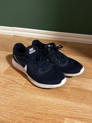 Marinblå Nike Revolution sneakers - Säljer ett par marinblå Nike Revolution sneakers i storlek 43. Skorna har vit sula, svart och vit yttersula samt vit Nike-logga på sidan. Ovandelen är i mesh-liknande syntetmaterial och de har klassisk snörning. Perfekta för träning eller vardagsstil.