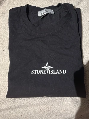 Svart Stone Island t-shirt med tryck storlek S - Svart t-shirt från Stone Island med stort logotryck på ryggen och mindre logga framtill. Klassisk rund hals och korta ärmar. Ikonisk patch på vänster ärm. Tillverkad i mjuk bomull för skön känsla och snygg streetwear-look. Storlek S