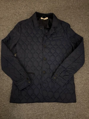 Mörkblå quiltad jacka Close by Denim - Säljer en mörkblå quiltad jacka från Close by Denim i storlek M. Jackan har klassisk krage, knäppning med knappar och två fickor framtill. Snyggt mönster i quiltad design och långärmad modell. Perfekt för dig som vill ha en stilren och bekväm jacka.