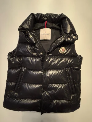 Svart dunväst från Moncler  - Säljer en svart, glansig dunväst från Moncler med hög krage och broderad logga på bröstet. Västen har dragkedja och knappar framtill samt två snedställda fickor med dragkedja. Den är 100% äkta och köptes för 270€ från MYTHERESA jag har också kvittot kvar från köpet så att skriv till mig om ni vill se det eller om ni har några andra frågor. Den är Max använd 5 gånger