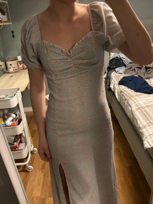 Ljusblå långklänning med blommönster - Supersöt långklänning i ljusblått med små orange blommor, puffärmar och en snygg slits framtill. Klänningen har en smickrande v-ringning med rynkdetalj och sitter figurnära upptill. Perfekt för sommarens alla tillfällen!