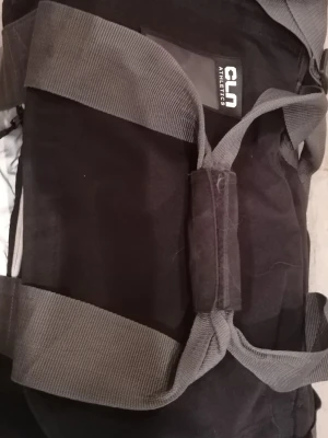 Svart träningsväska CLN Athletics - Säljer en svart träningsväska från CLN Athletics med stora vita logotexter på sidorna. Väskan har breda grå axelband och ett vadderat handtag för extra komfort. Tillverkad i slitstark polyester och perfekt för gymmet eller sportaktiviteter.