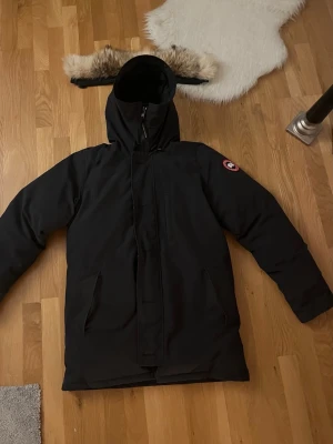 Svart dunjacka från Canada Goose - Säljer en svart dunjacka från Canada Goose med avtagbar huva och fuskpälsdetalj. Jackan har dragkedja framtill, två stora fickor och klassisk patch på ärmen. EN liten defekt vid armhålan (skriv privat). 100% äkta har tyvärr inget kvitto. Var öppen med funderingar! 