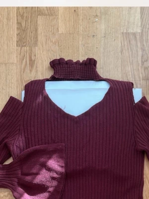 Tröja  - Vinröd ribbad polotröja med hög krage och v-ringning. Tröjan har långa ärmar med breda trumpetärmar som ger en unik look. Materialet är stretchigt och stickat, perfekt för höst och vinter. Snygg detalj med cut out vid axlarna.