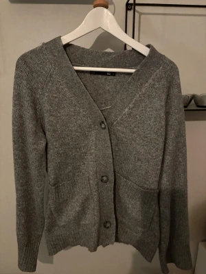 Grå stickad kofta med knappar - Mysig grå stickad kofta  med v-ringning och stora knappar framtill. Koftan har långa ärmar, två fickor och ribbade muddar vid ärmslut och nederkant. Perfekt att slänga på sig när det är kyligt ute.