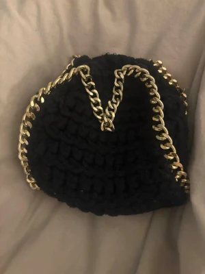 Svart stickad axelväska med kedja - Säljer en svart stickad axelväska med grov guldfärgad kedja runt kanterna och som handtag. Väskan har även sopiga silverfärgade kedjor för axelrem. Perfekt storlek för det viktigaste och unik look med chunky stickat material.