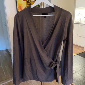 Brun omlottkofta från H&M - Säljer en brun omlottkofta från H&M i storlek S. Koftan har långa ärmar, knyt i sidan och är tillverkad i ett tunt, mjukt material som känns skönt mot huden. Perfekt för lager på lager och enkel att styla till olika looks.