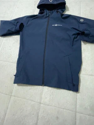 Marinblå vindjacka från Sail Racing - Säljer en marinblå vindjacka från Sail Racing med huva och hel dragkedja framtill. Jackan har diskret logga på bröstet och en rund detalj på ärmen. Tillverkad i slitstarkt syntetmaterial, perfekt för blåsiga dagar.